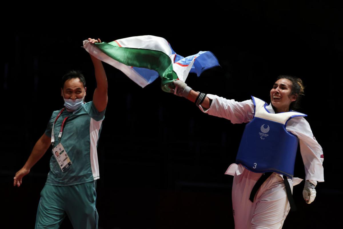 A smiling male coach and female athlete wave an Uzbek flag between them after a competition bout.