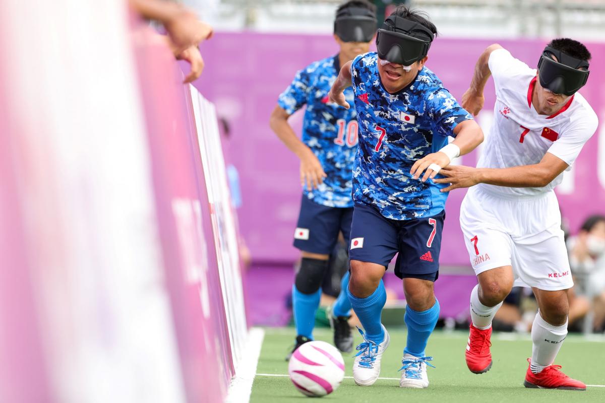 A blind football player in a blue jersey competes for the ball against a player in a white jersey at Tokyo 2020 Paralympic Games.
