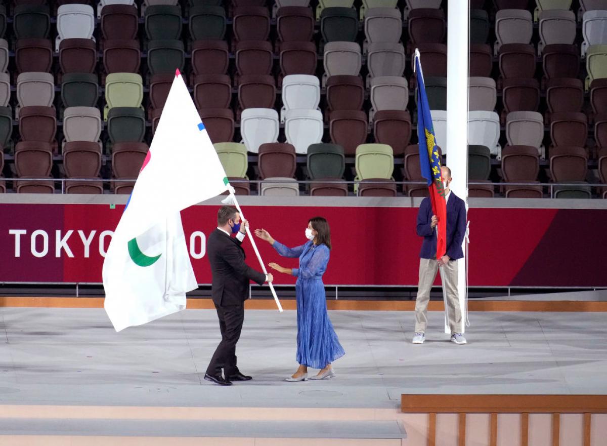 A man passes the Paralympic flag to a woman who stretches out her hands to receive it.