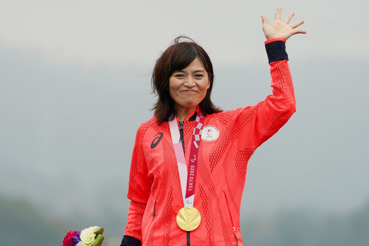 A Japanese female athlete with a gold medal around her neck waves her hand.