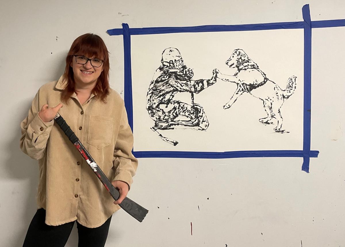 A female artist holding a Para ice hockey stick and stands in front of a painting of a female Para ice hockey player and her dog.