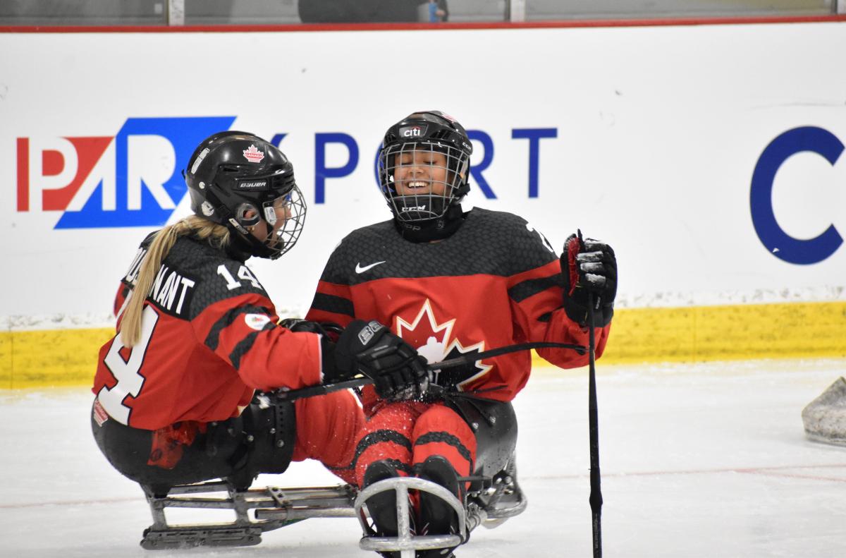 Two female Para ice hockey players on a sled smiling under their helmets in red jerseys.