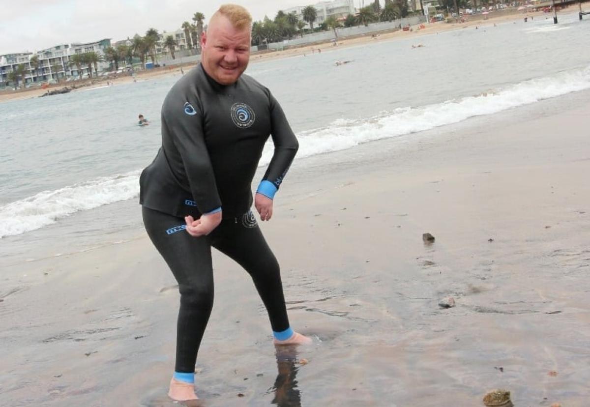 A male in a wet suit stands on the ocean shore, his feet in the water.
