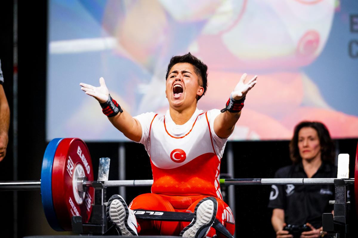 A female powerlifter raises her hands and yells in celebration while seated on a bench.