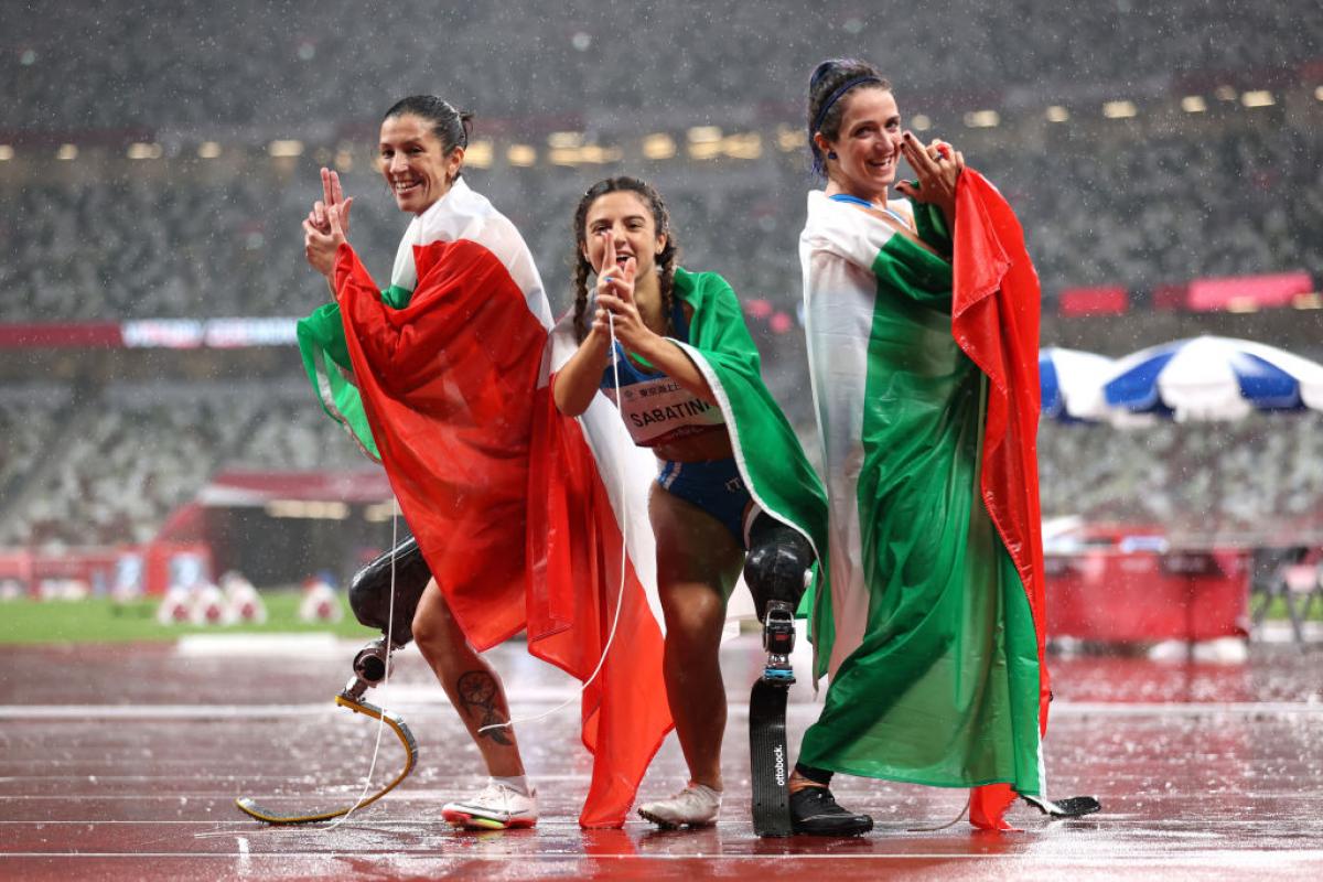 Three female athletes pose by raising two fingers in front of their faces at the Tokyo 2020 Paralympic Games.