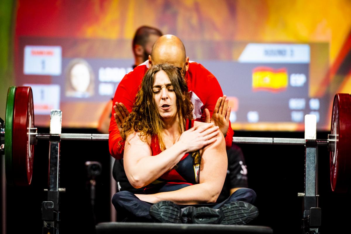 A female powerlifter on the bench as her coach gives her a last minute shoulder warm-up.