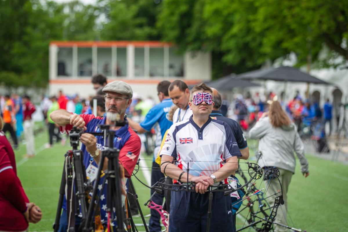 An archer in a blindfold decorated with the Union Jack rests his hands on a downturned bow.
