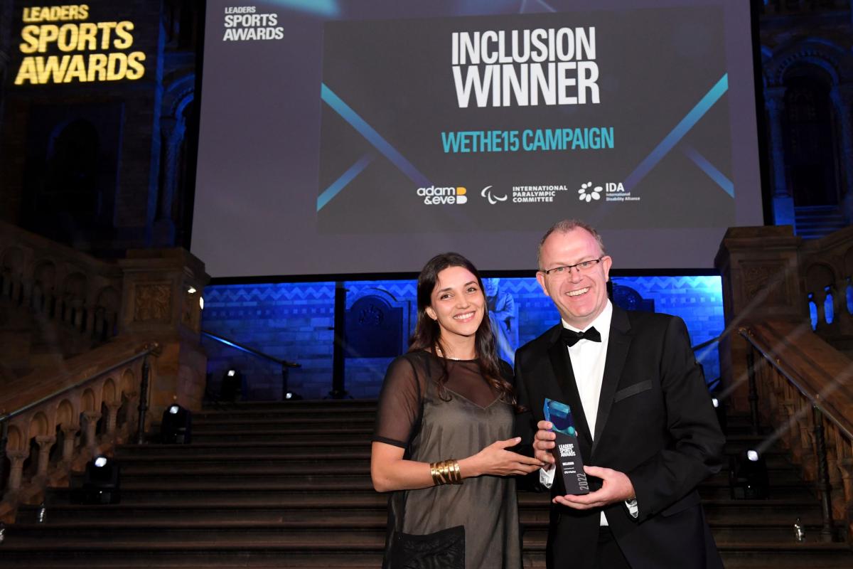 A man and a woman hold a trophy in front of a screen with the words "Inclusion Winner WeThe15 Campaign"