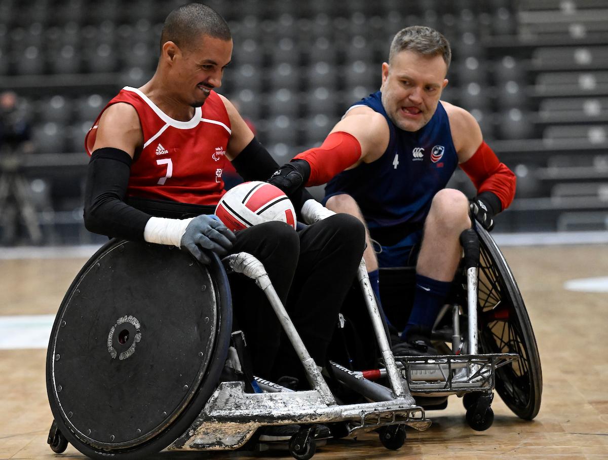 A male wheelchair rugby player wearing a red jersey carries the ball, while another player reaches for the ball.