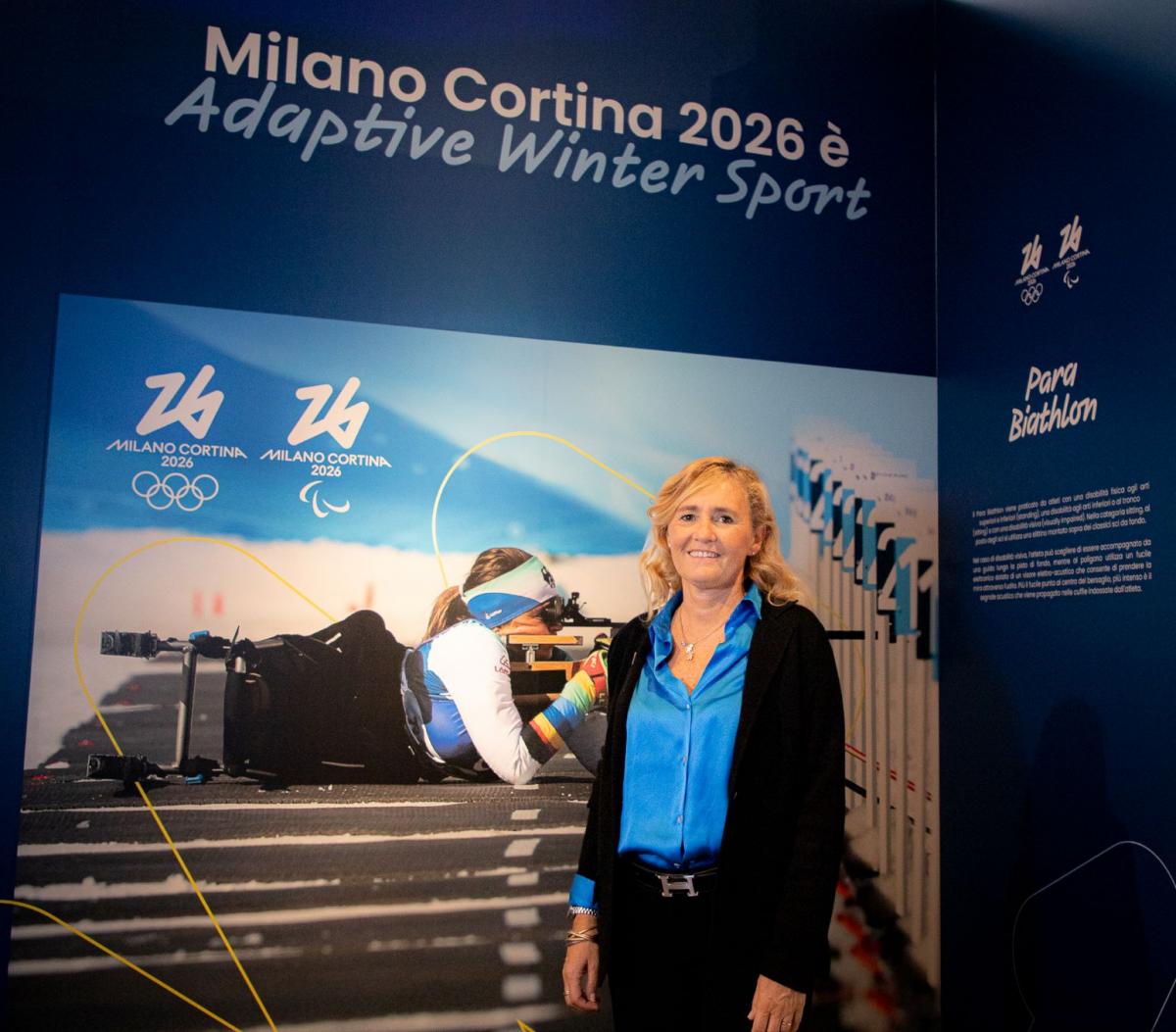 A woman in business attire poses in front of a poster featuring a Para biathlon athlete in action.