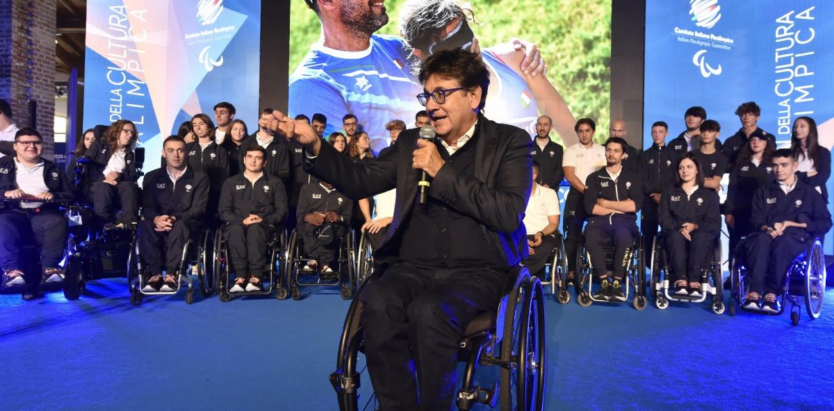 A man in a wheelchair speaks in a microphone on stage with a crowd of athletes behind him.
