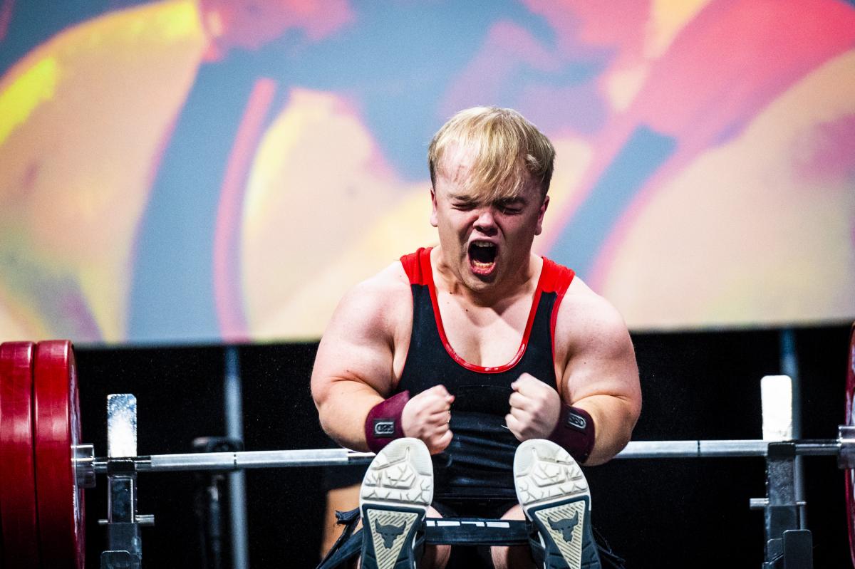 A male Para powerlifter of short stature screams and squeezes his fists in celebration after making a successful lift.