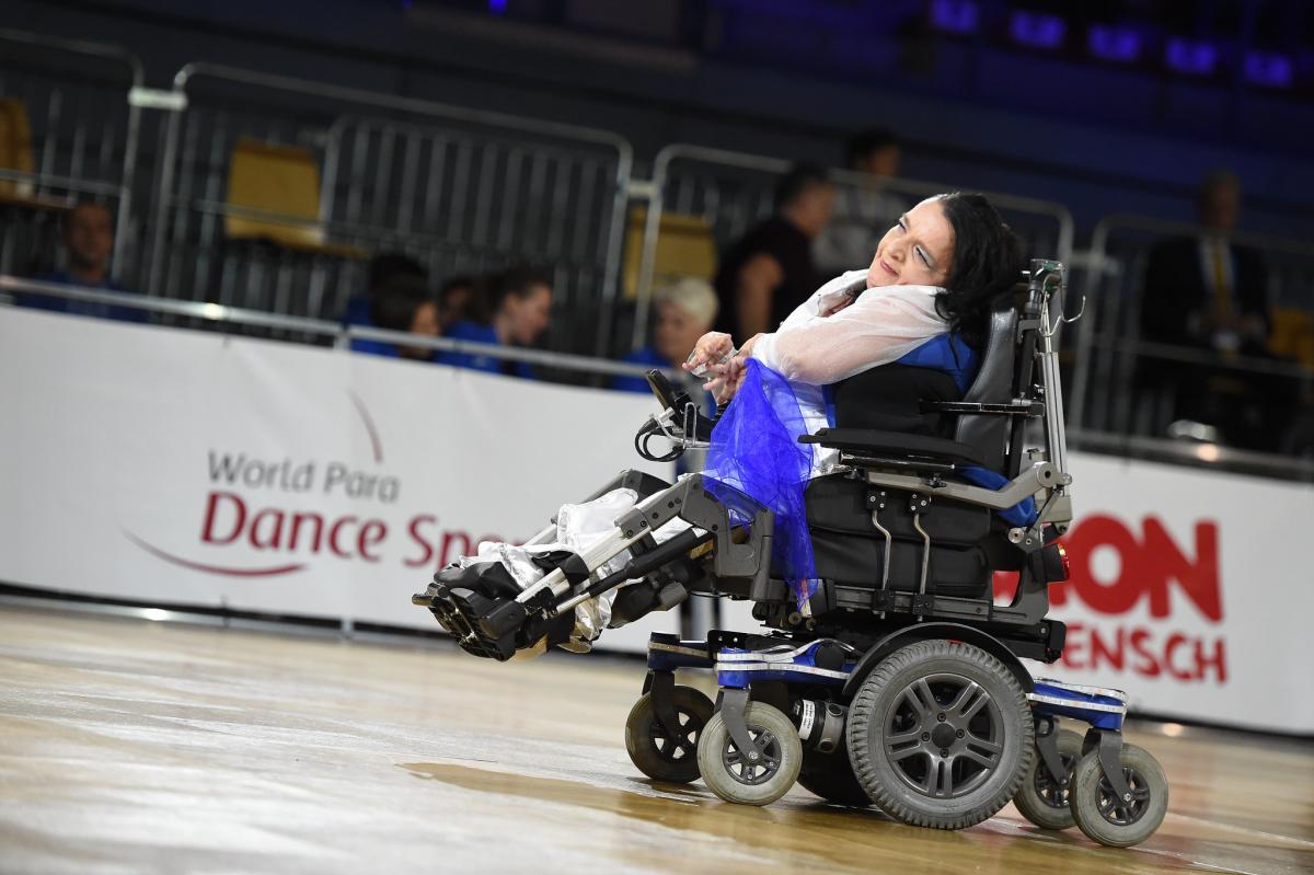 A female dancer in an electric wheelchair smiles and motions with her hands during a dance performance.