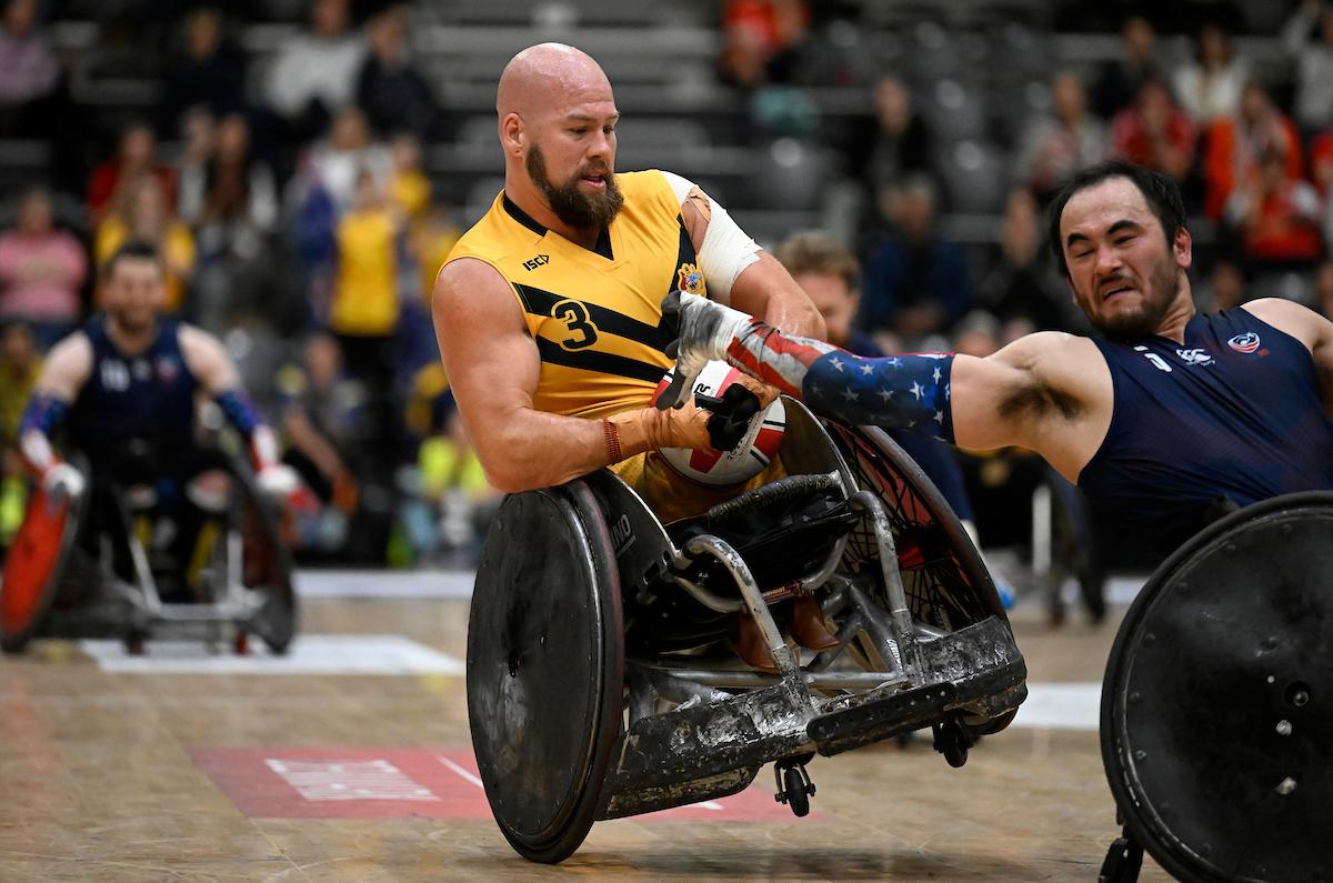 A male wheelchair rugby player wearing a yellow jersey holds onto a ball, while another athlete wearing a blue jersey tries to grab it