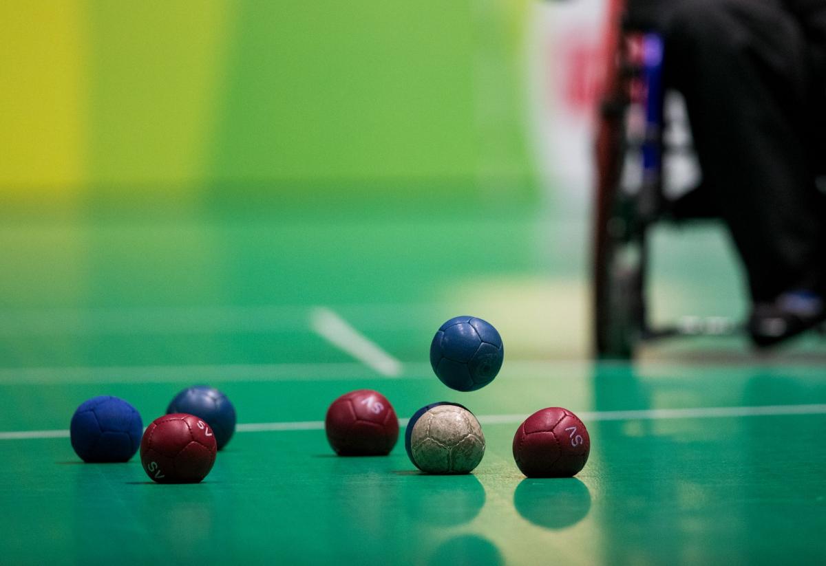 Seven boccia balls lying on the playing ground while one blue ball is falling down, about to touch the ground.