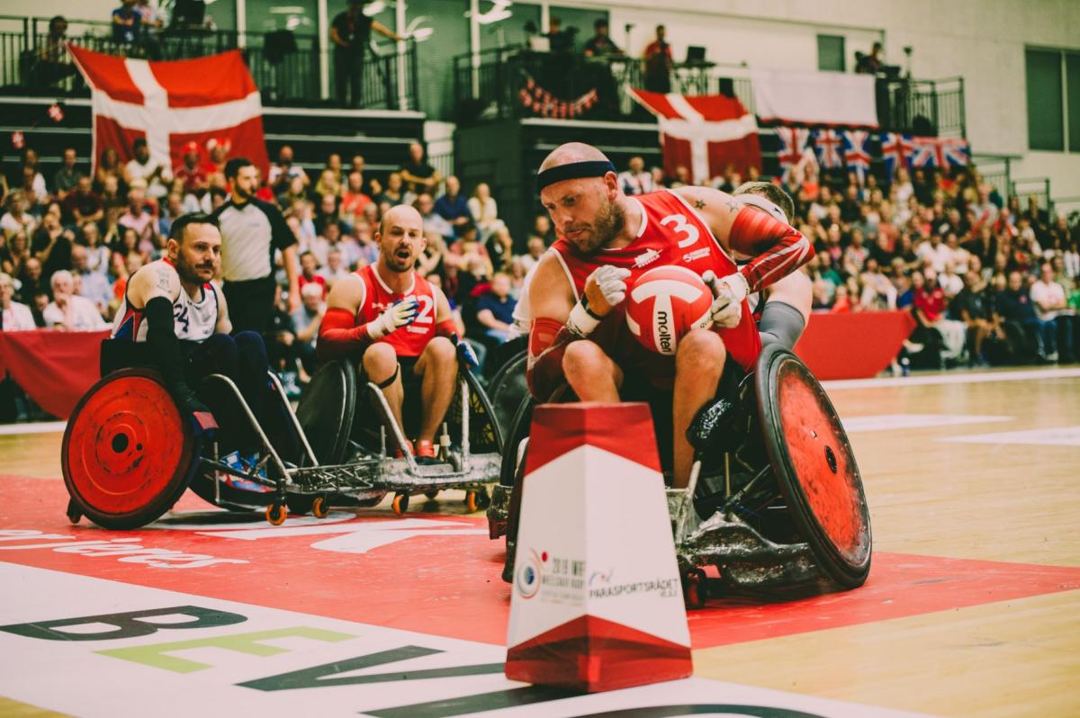 Three male athletes in wheelchairs, with one carrying a red and white ball, play at a venue packed with spectators.