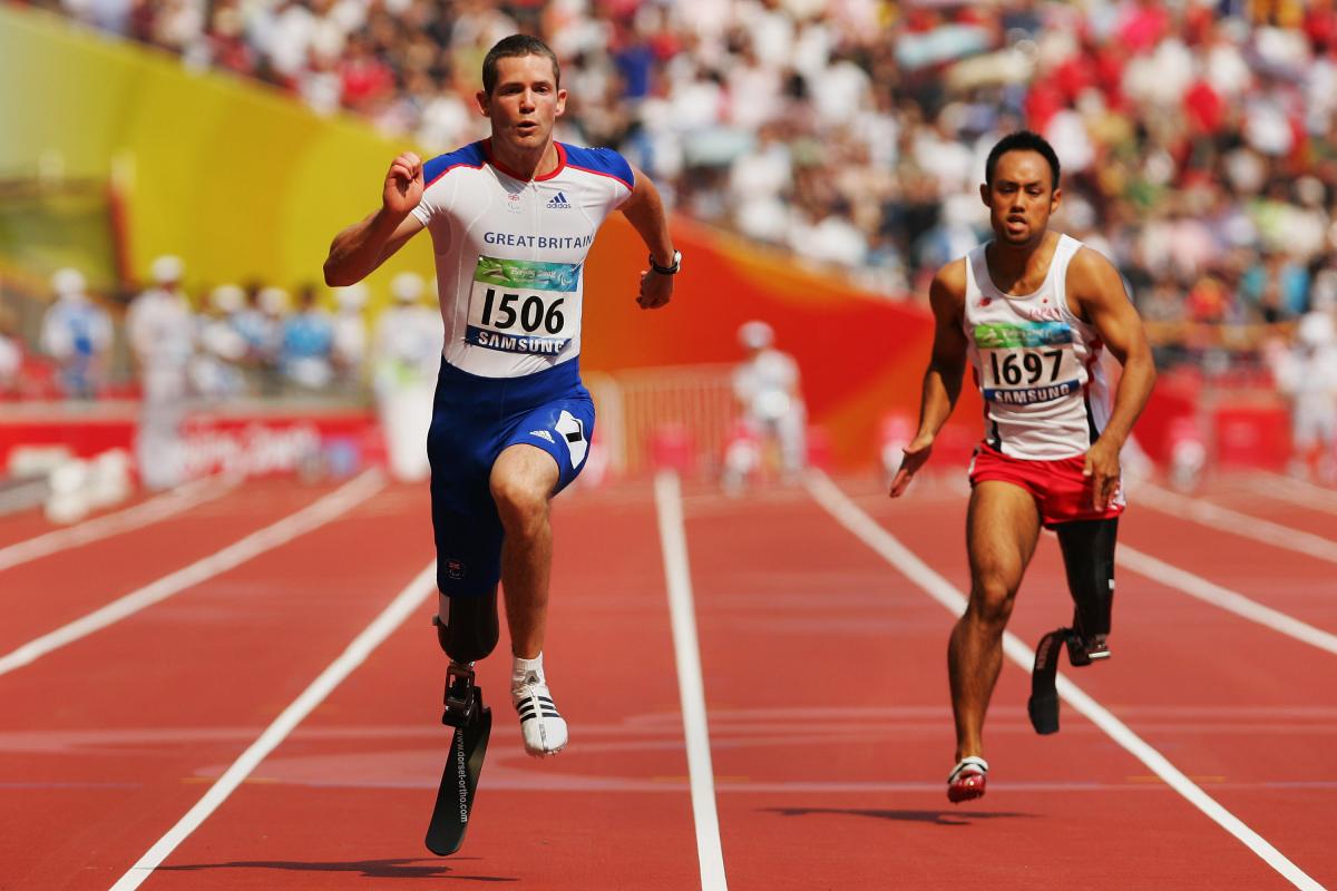Two men with prosthetic legs sprint on an athletics track at a packed stadium.