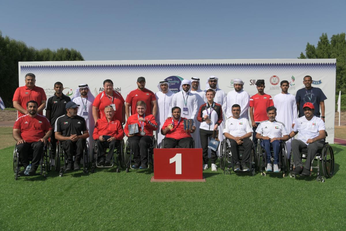 A group of nine men in wheelchairs posing in a podium with 14 people behind them