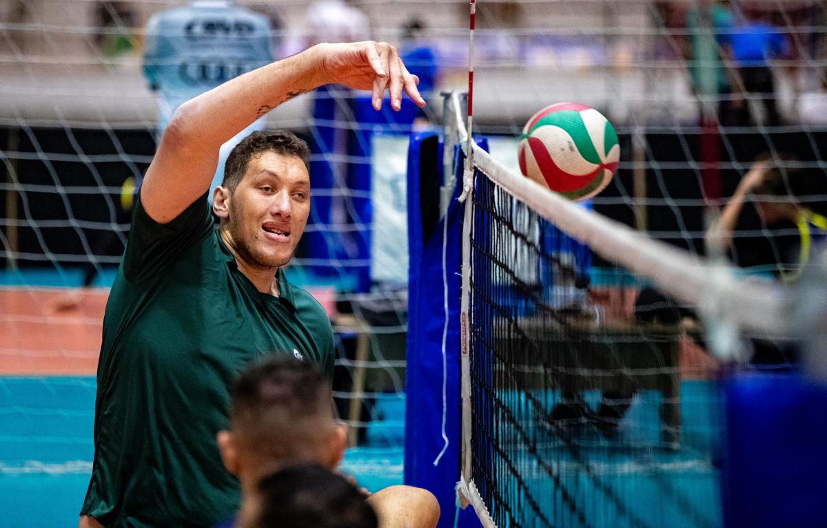 A male sitting volleyball player raises his right arm to hit the ball above the net