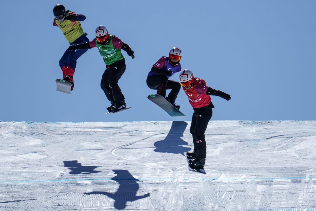 Four male snowboarders, all wearing vests that say Beijing 2022, helmets and goggles, compete at the Beijing 2022 Paralympic Games