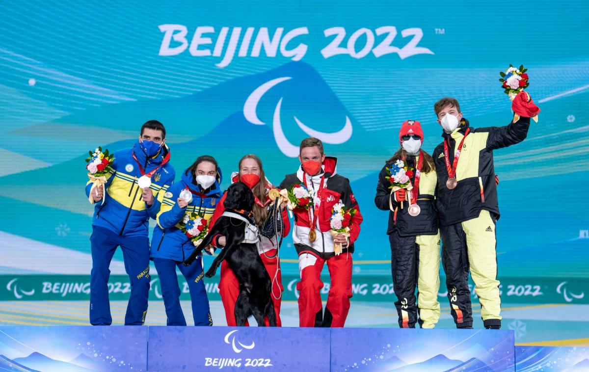 Six athletes pose for a photograph on the podium. A female athlete holds a gold medal with her left hand and her black guide dog with her right hand.