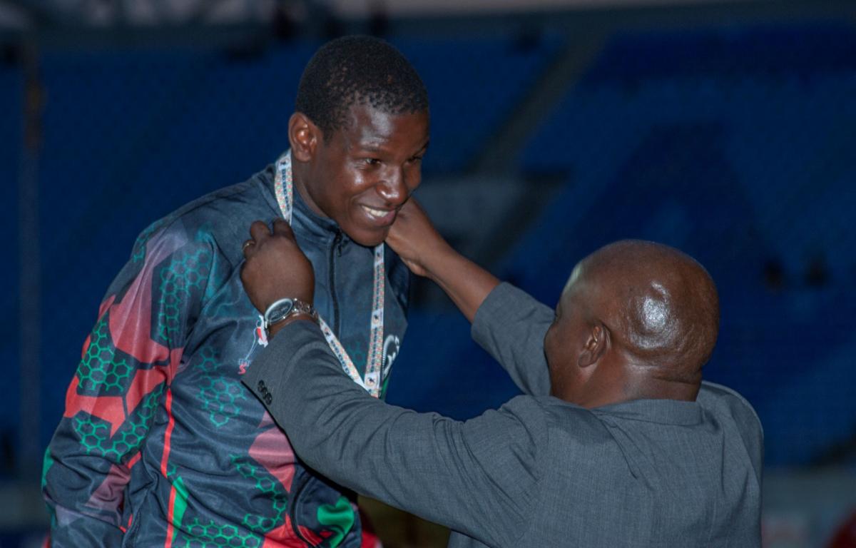 A male athlete in a national kit smiles as a medal presenter puts a medal around his neck.