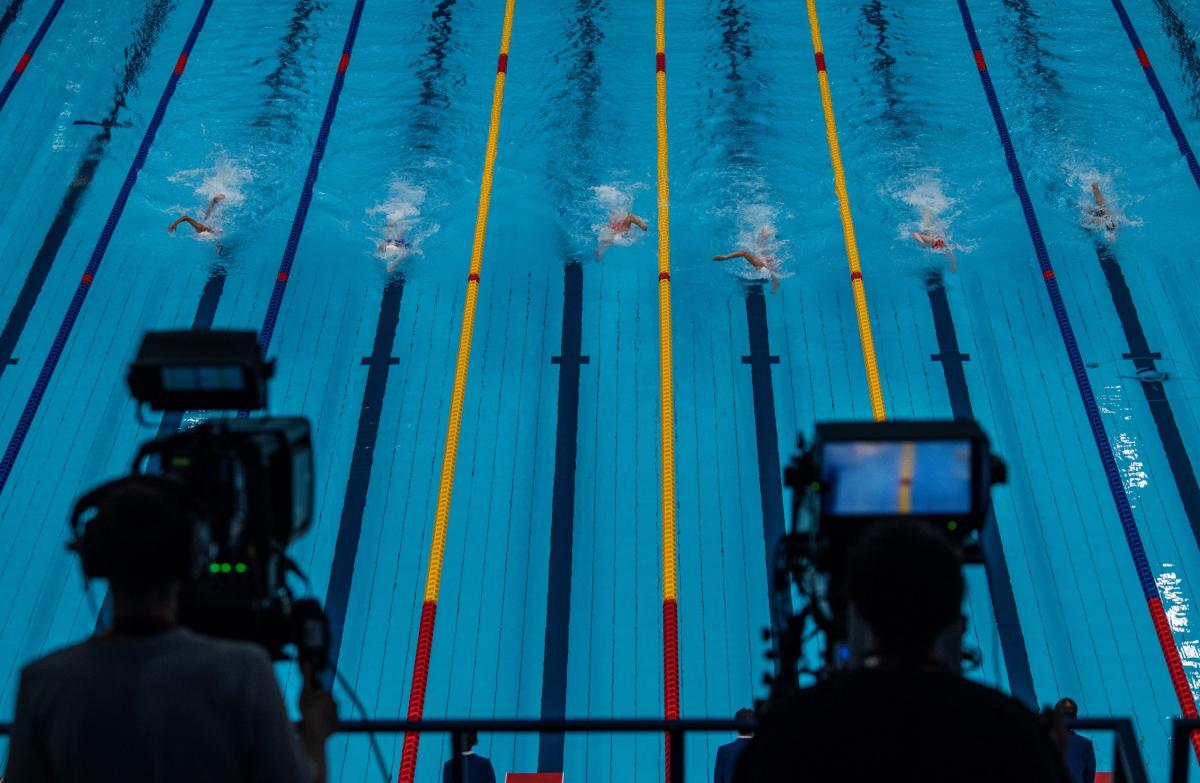 A look at a competition pool in Tokyo, with six swimmers in the lanes, through two cameras that are filming the race.