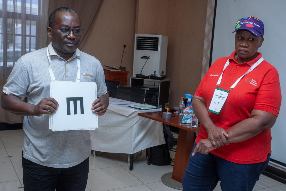 A man holds up charts for testing eyesight while a female supervisor looks on.