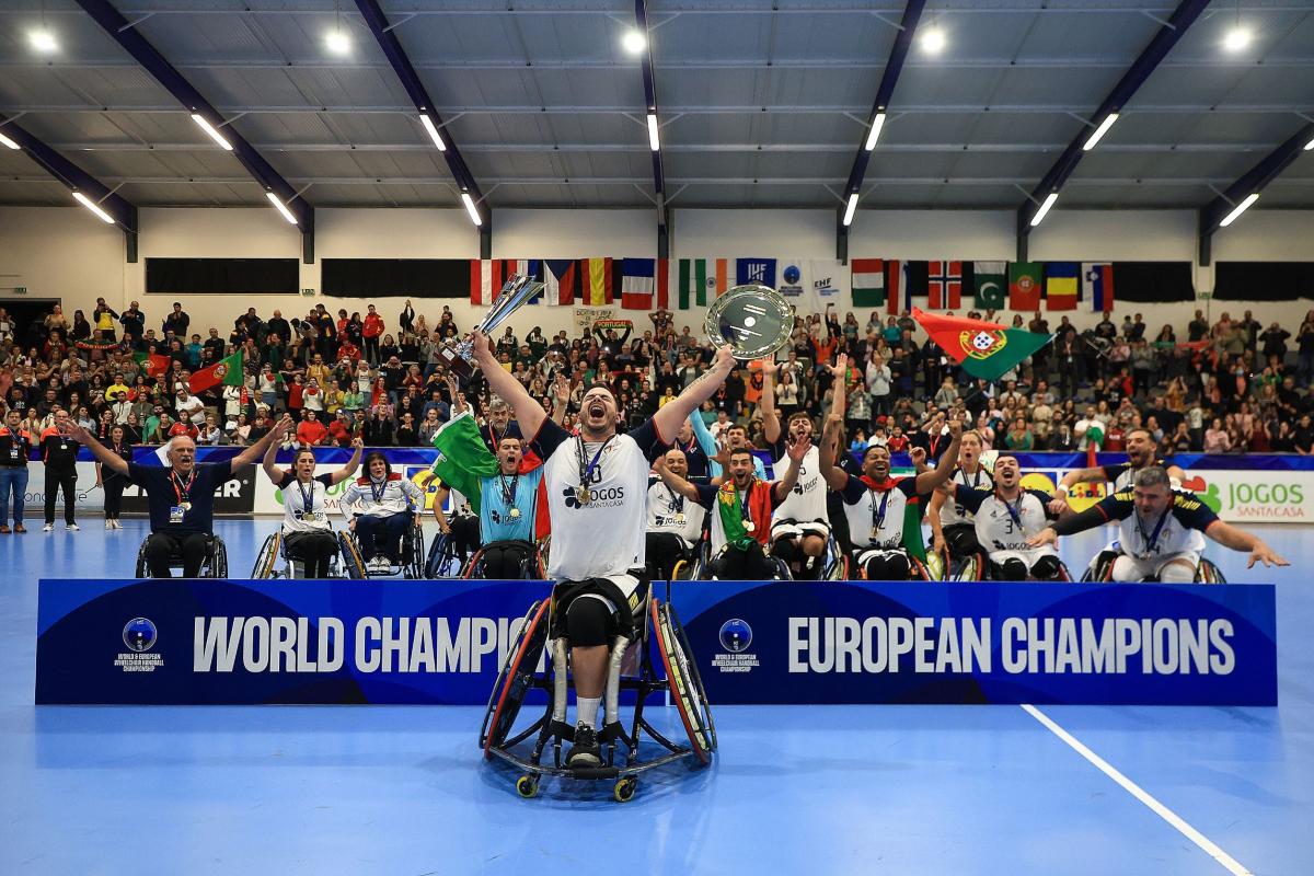 A male athlete in a wheelchair screams in celebration while holding up a trophy and dish, as a team cheers behind him next to the banner "world champions" and "European champions".