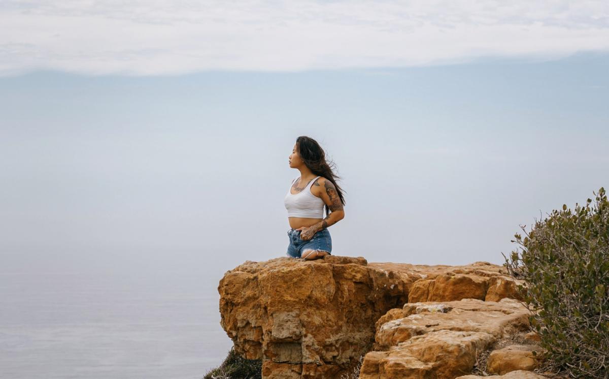 A young female without legs below the hips stands on a cliff looking out a large body of water.