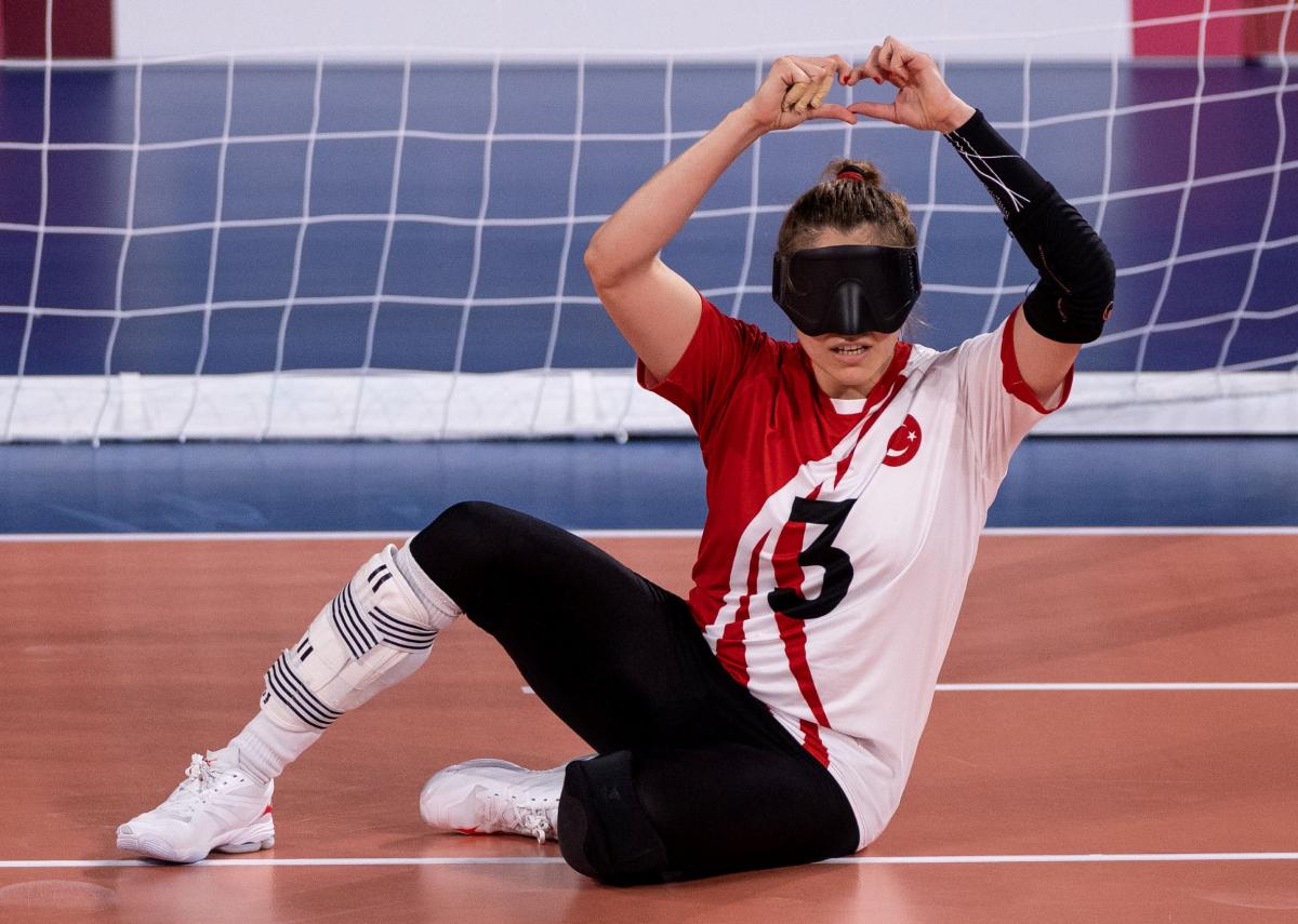 A female goalball player in an eye mask makes a heart gesture with her hands as she sits on the competition field.