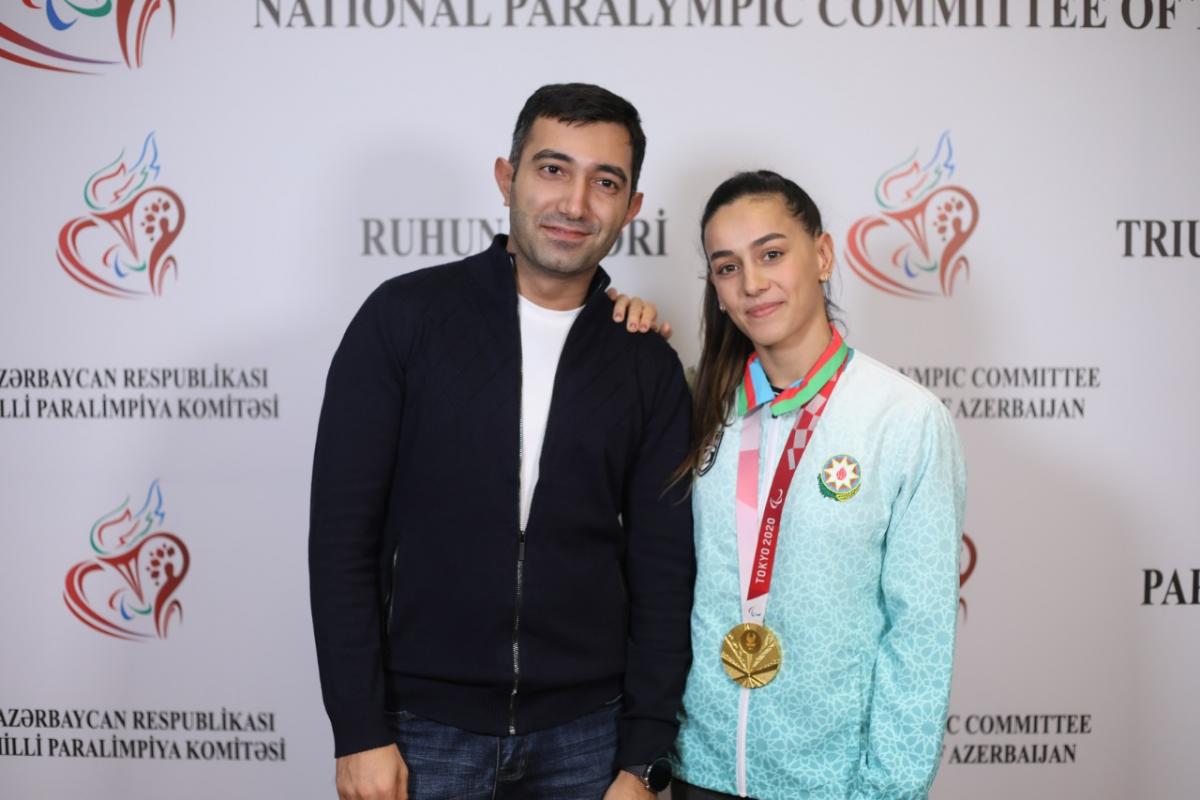 A man in and a woman, her right hand on his shoulder and a gold medal around her neck, pose in front of an official backdrop.