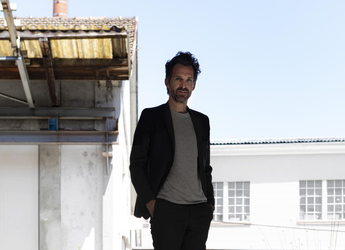 A man in a T-shirt and black suit poses with the backdrop of a slightly dilapidated house.