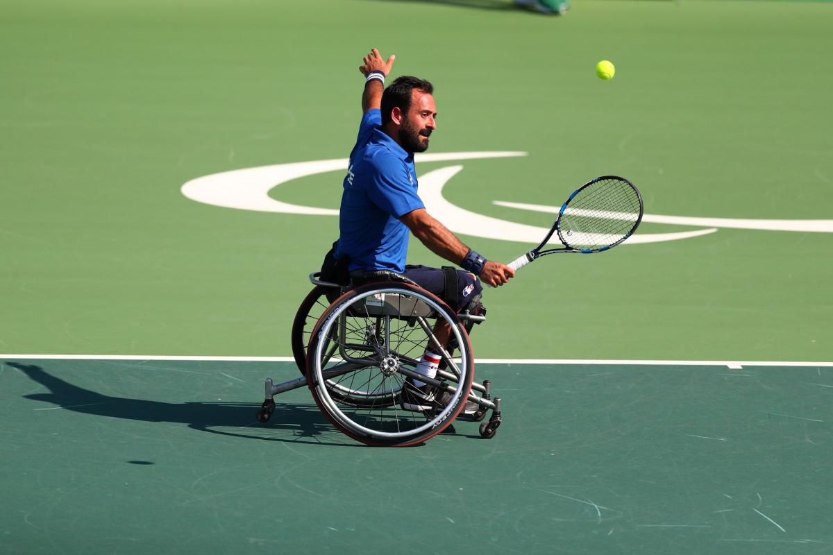 A male wheelchair tennis player makes a serve on a hard green court with his left arm stretched out wide.