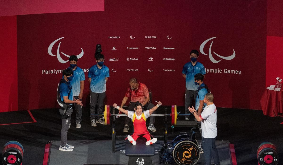 A female Para powerlifter celebrated on stage with five people around her