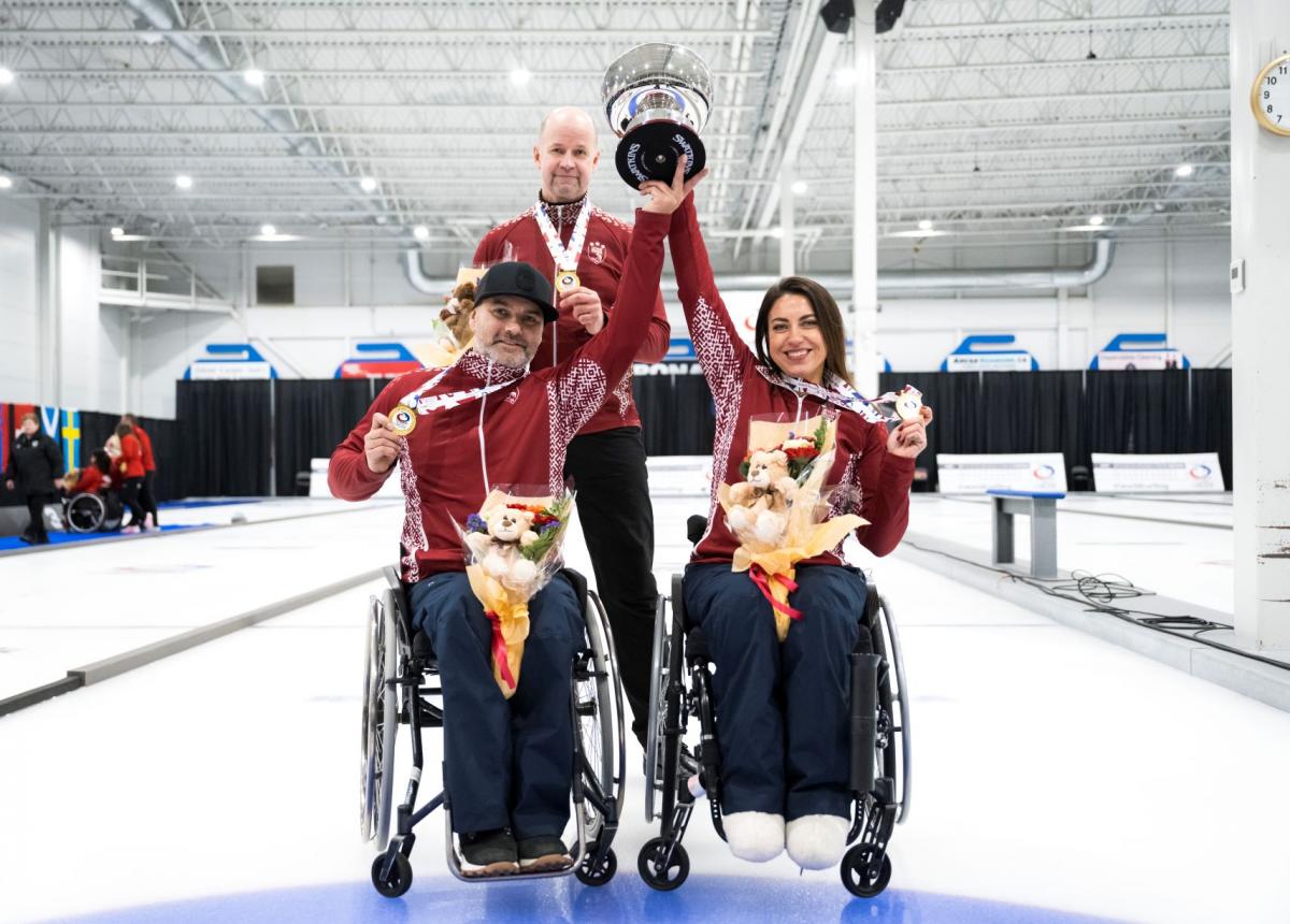 A female wheelchair curler and a male wheelchair curler lift a trophy together, while showing a gold medal