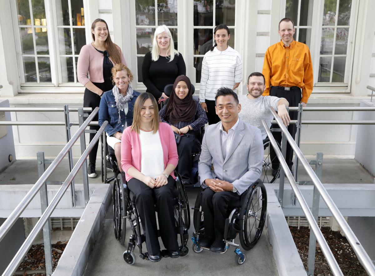 Nine athletes, five in wheelchairs at the front and second rows, and four standing in the back pose for a photo in front of the IPC headquarters.