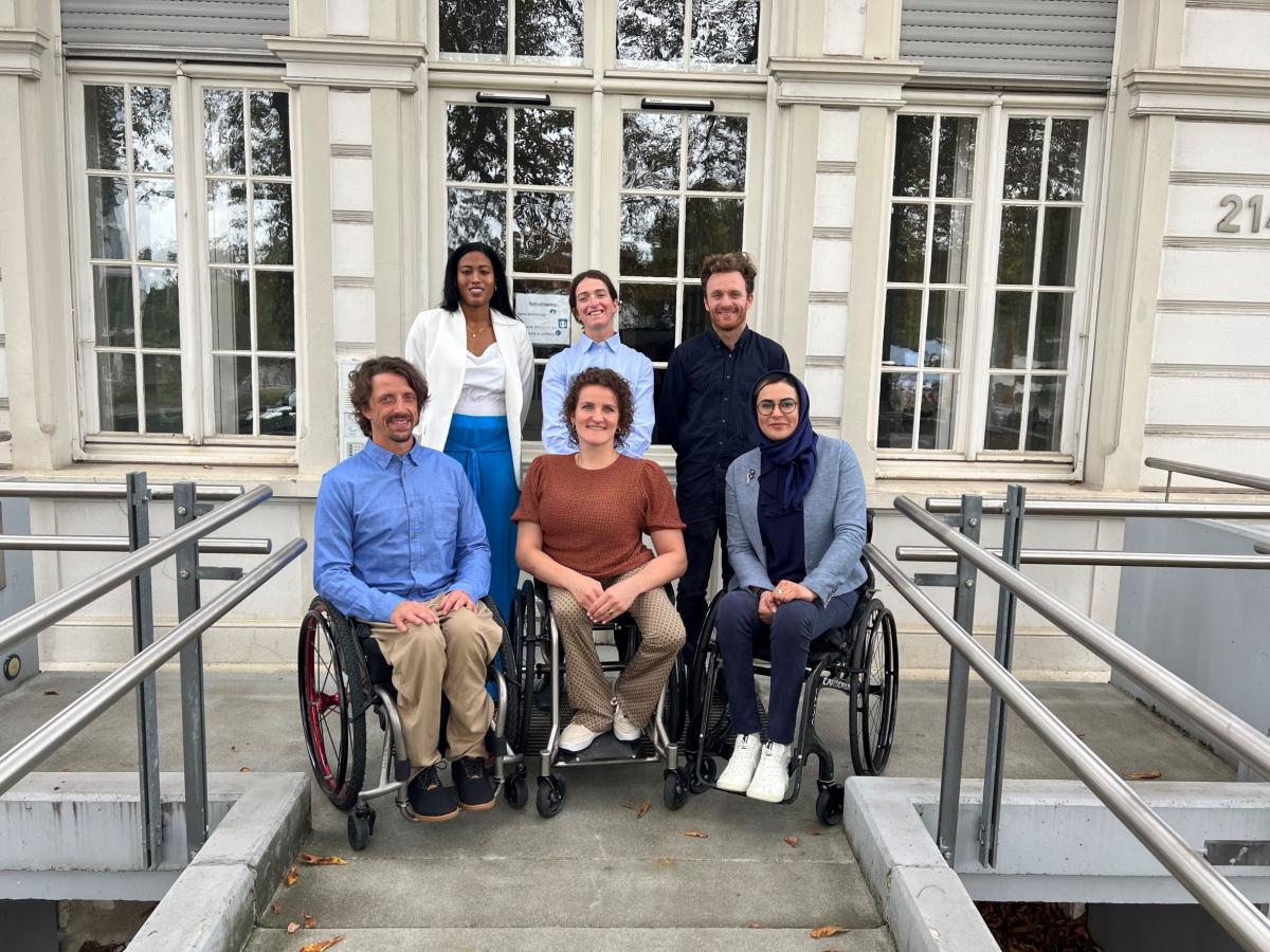 Six athletes, three standing in the back and three in wheelchairs at the front, pose for a photo in front of the IPC headquarters.