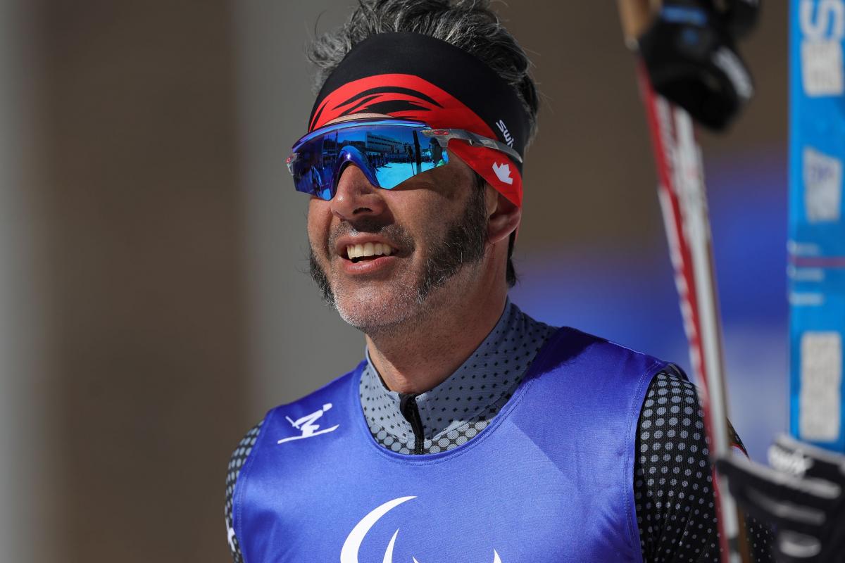 A close-up of a male cross-country skier in sunglasses and competition bib, holding skis in one hand. 