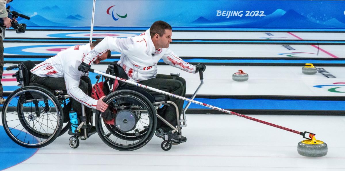 A male athlete slides a curling stone using a stick on the ice at Beijing 2022. He is on a wheelchair and another male athlete is holding onto the tires.