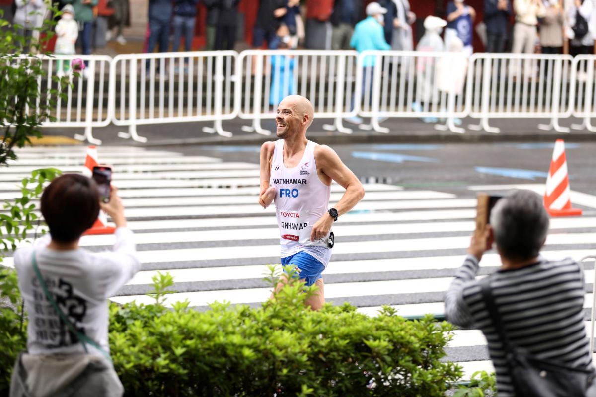 An athlete who is missing his right arm below the elbow smiles as he runs on the street during a marathon as onlookers take photos of him.