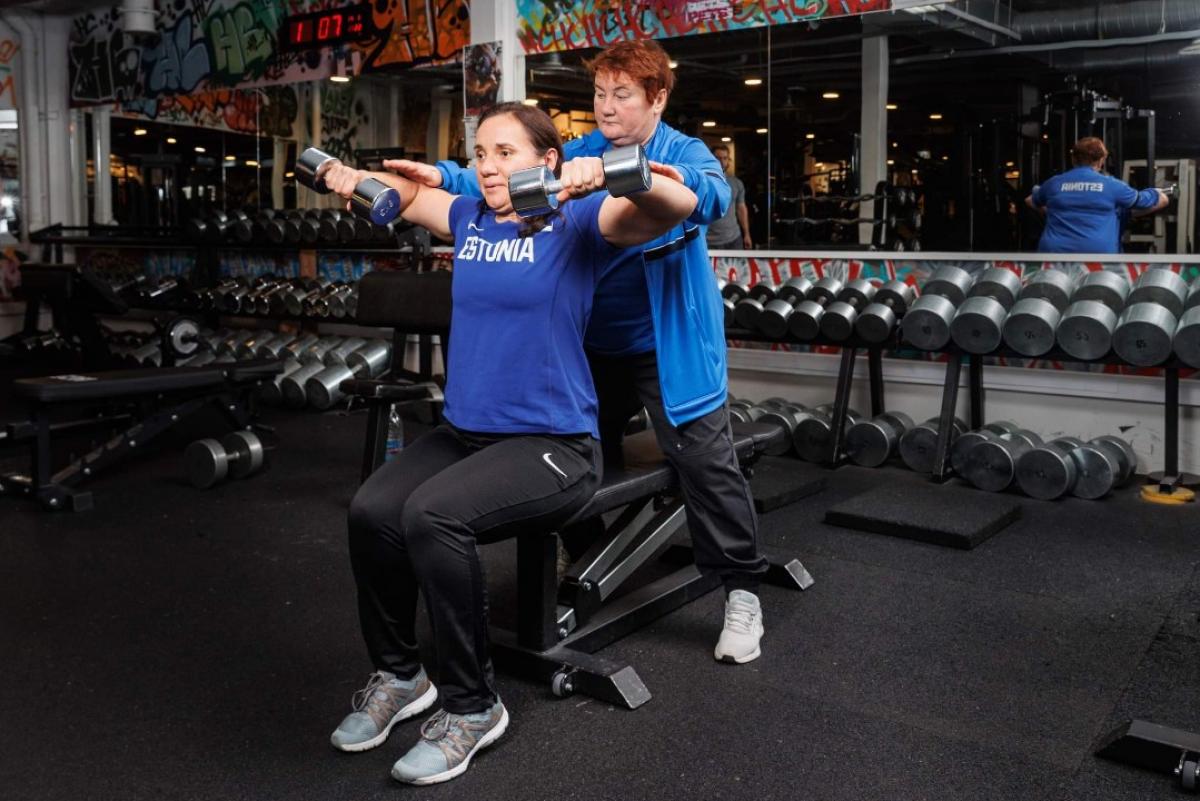 A female athlete in an Estonian T-shirt lifts two dumbbells in a gym as an older woman spots her from behind.