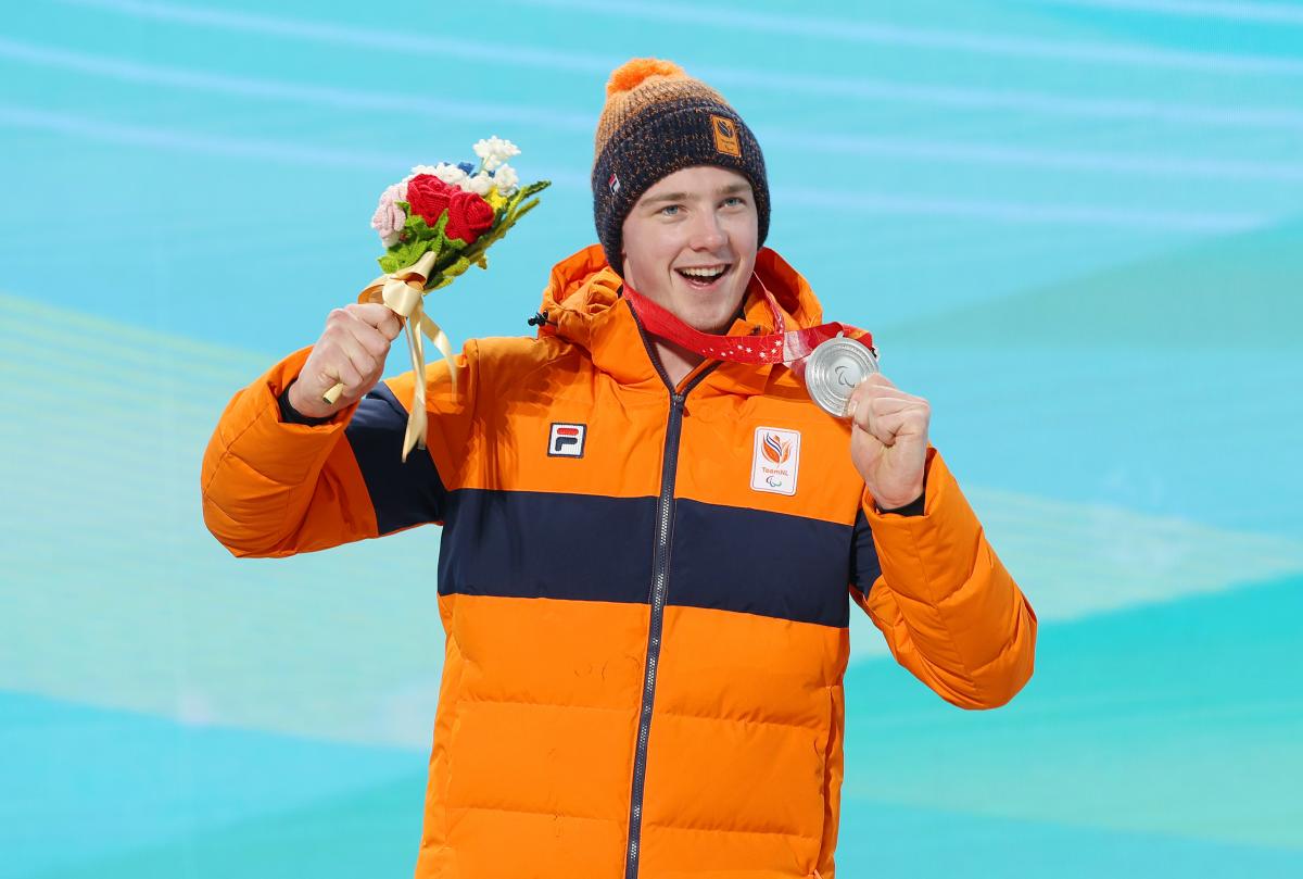 A smiling male athlete holds up a silver medal and bouquet of flowers during a medal ceremony. 
