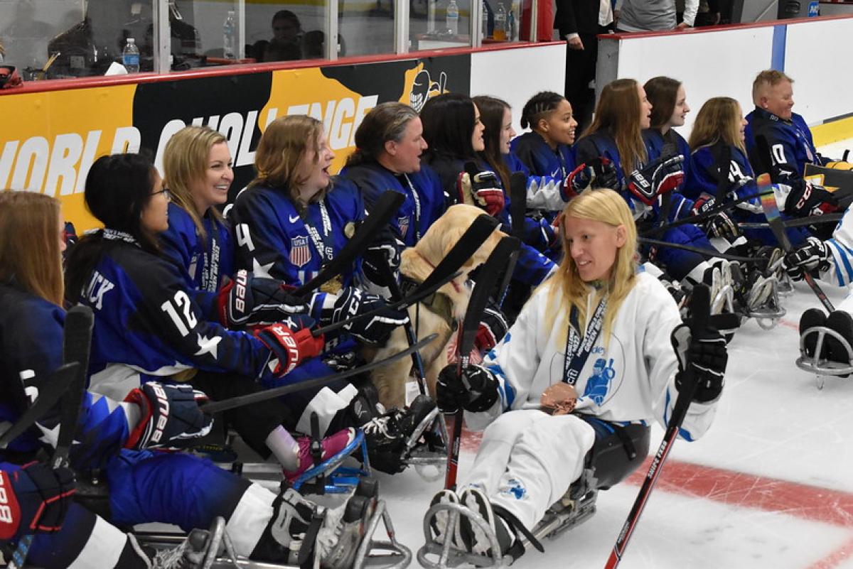 Female athletes in blue jersey are on the ice, while a female athlete in white jersey passes by.