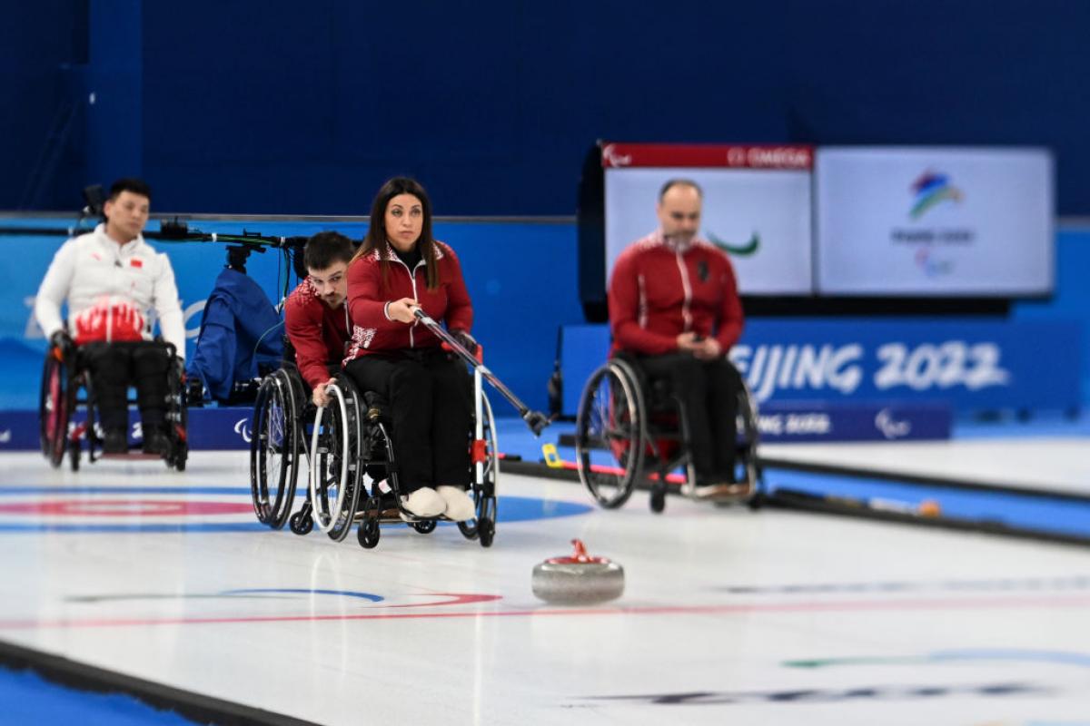 A female wheelchair curler delivers a stone with a stick, while a male athlete holds the wheels of her wheelchair.