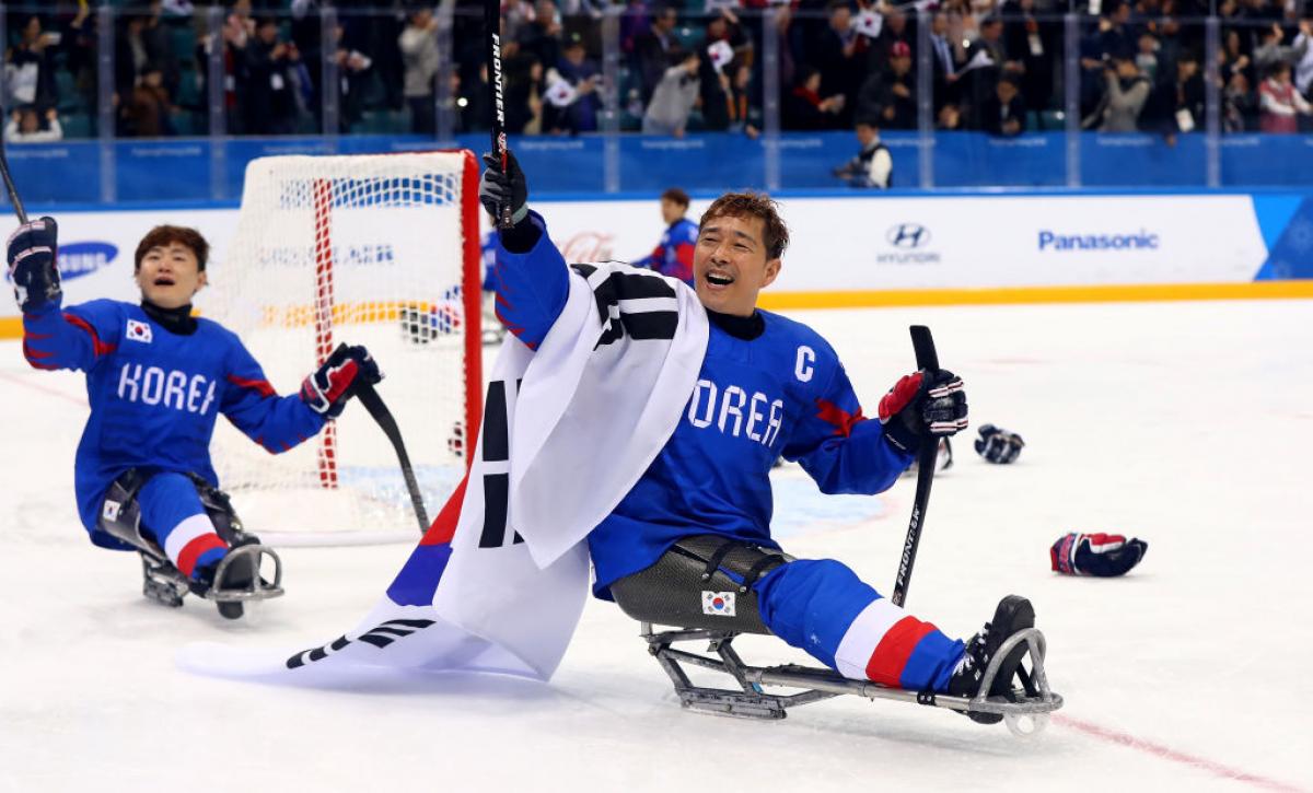 A male Para ice hockey player holds up a sledge hockey stick with his right hand and carries the Republic of Korea flag.