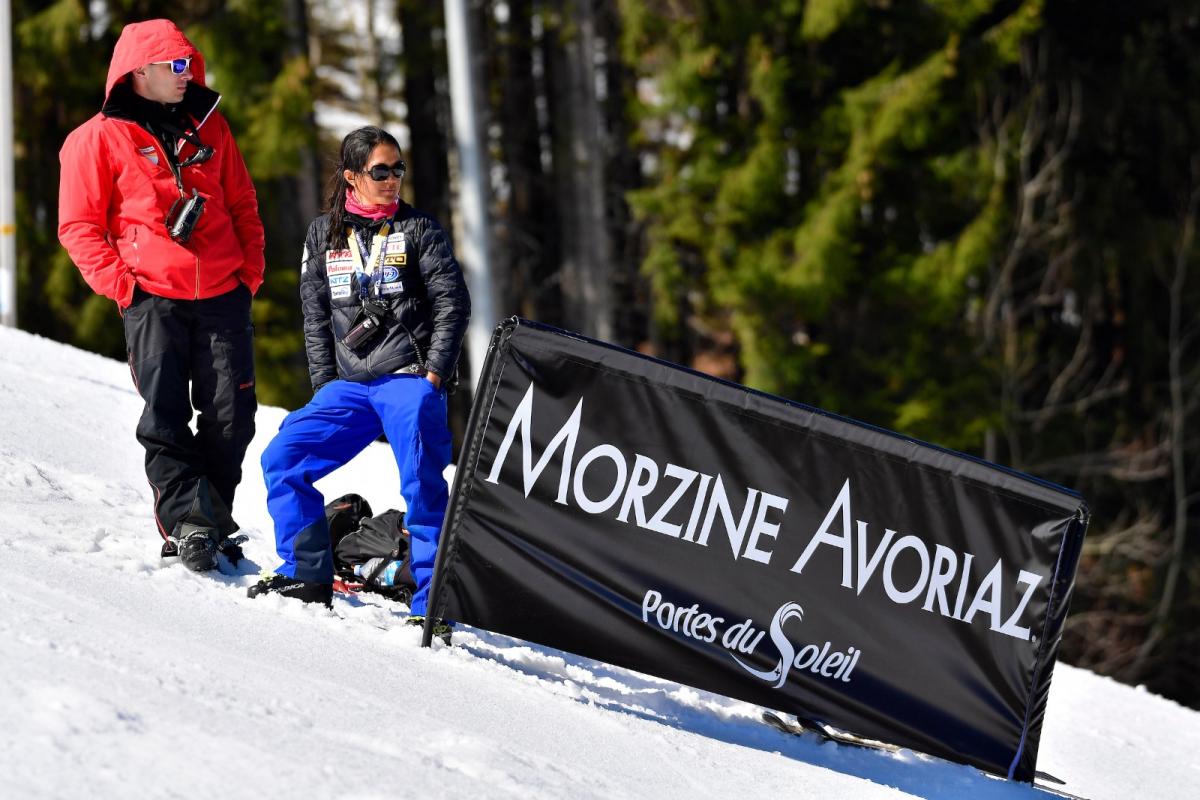 A female coach stands on the snow, next to a male staff and a sign that reads "Morzine Avoriaz"