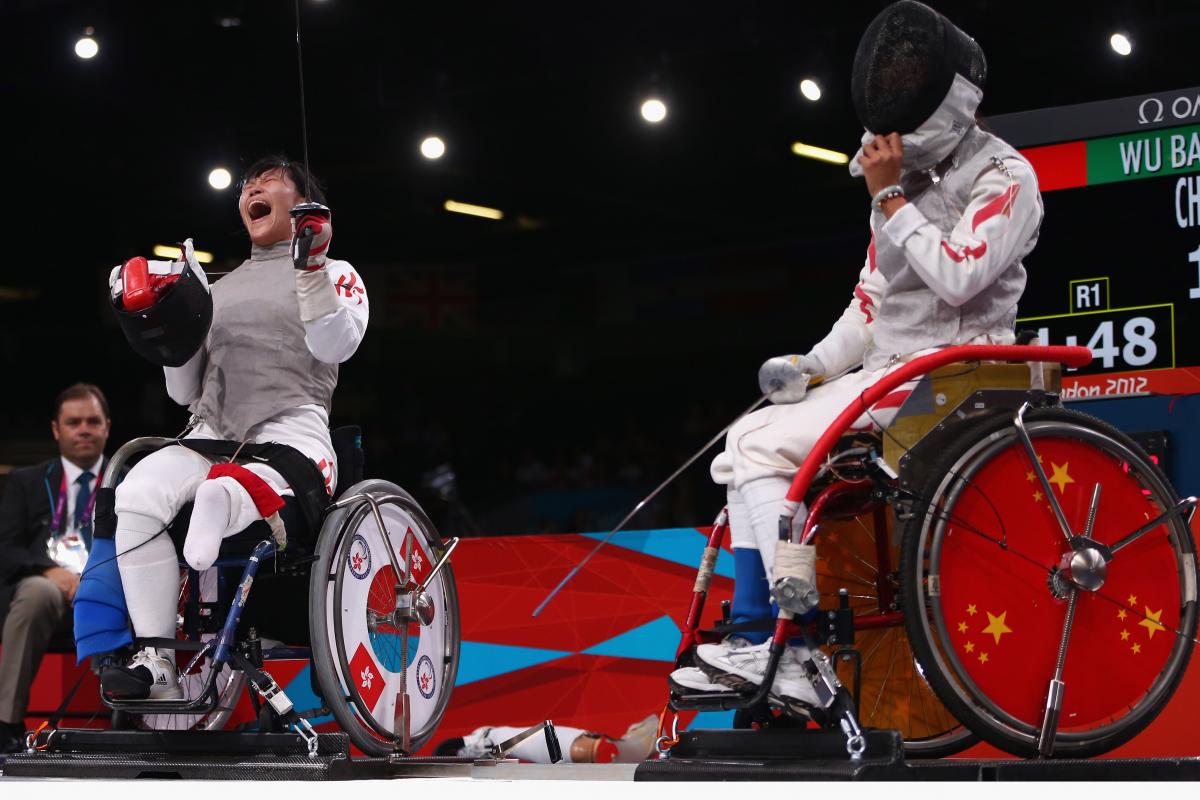 A female wheelchair fencer celebrates by taking her mask off with a shout after a winning bout.