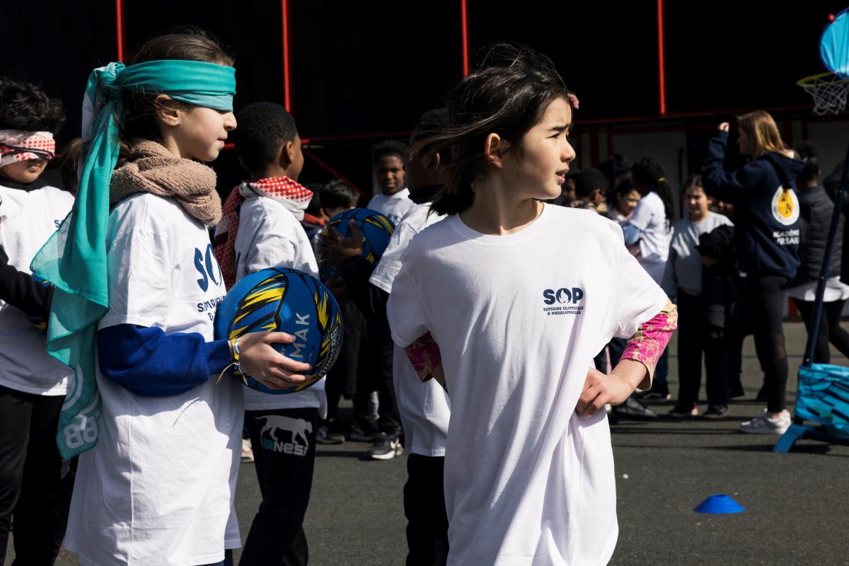 Two young girls, one with a blindfold, holding a ball, and the other looking back, participate in an event during Olympic and Paralympic Week.