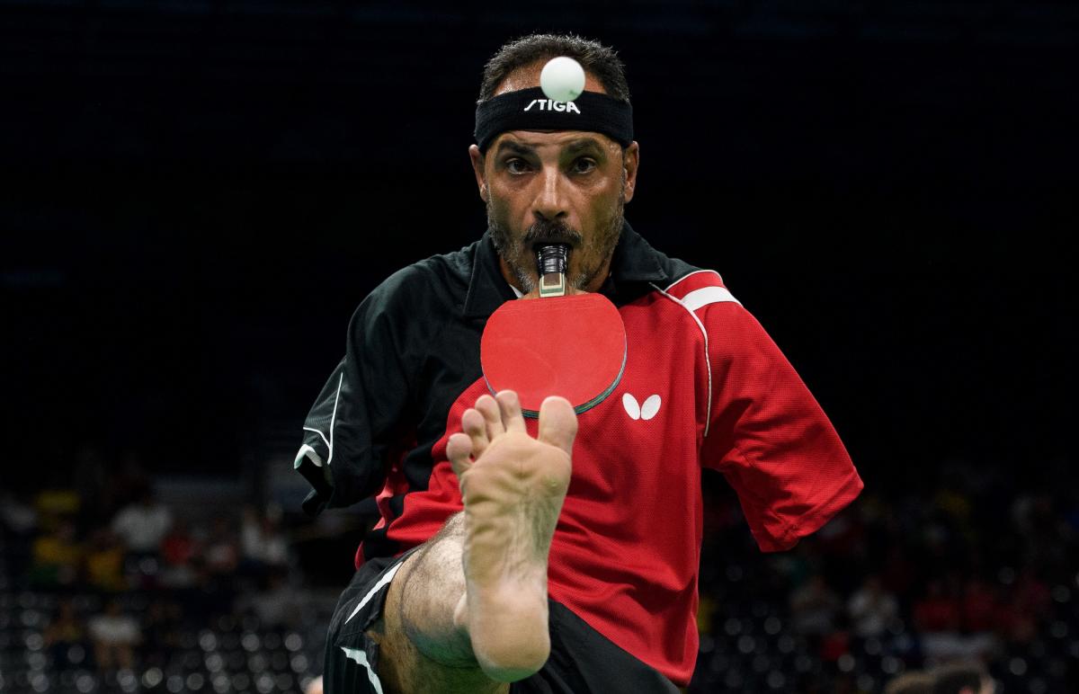 A male Para table tennis player holds a racquet with his mouth and lifts the ball with his right foot.